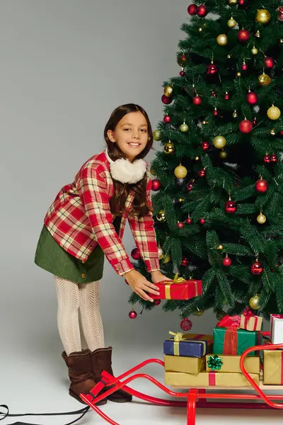 Cheerful girl places a gift under the sparkling Christmas tree with joy and excitement. — Stock Photo