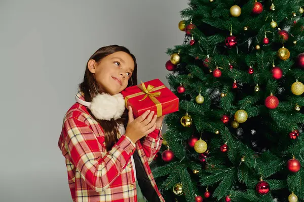Girl with a warm smile holds a red gift by a colorful, ornament filled Christmas tree. — Stock Photo