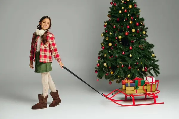 Happy girl wearing cozy attire drags a colorful sleigh filled with gifts by a Christmas tree. — Stock Photo