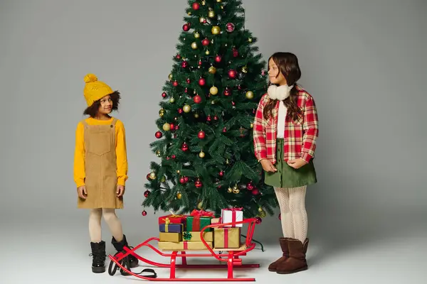 Two cheerful girls share laughter beside a festive Christmas tree with gifts nearby. — Stock Photo