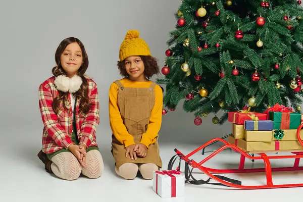 Two happy girls dressed in festive clothing sit beside a decorated Christmas tree with presents. — Stock Photo