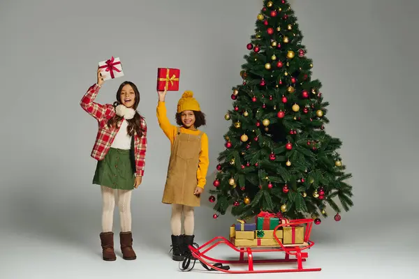 Two happy girls hold up brightly wrapped gifts beside a beautifully decorated Christmas tree. — Stock Photo