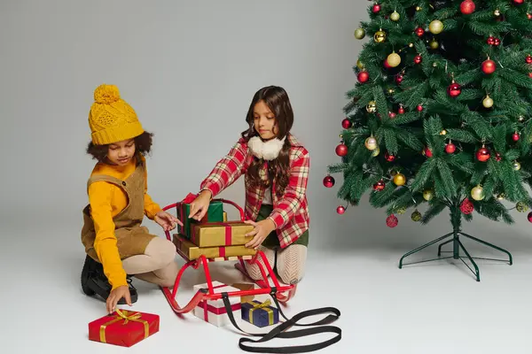 Two little friends excitedly arrange presents on a festive sleigh near a decorated Christmas tree. — Stock Photo