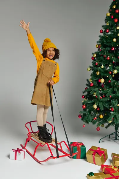 Girl joyfully stands on a sled surrounded by gifts and a festive Christmas tree. — Stock Photo