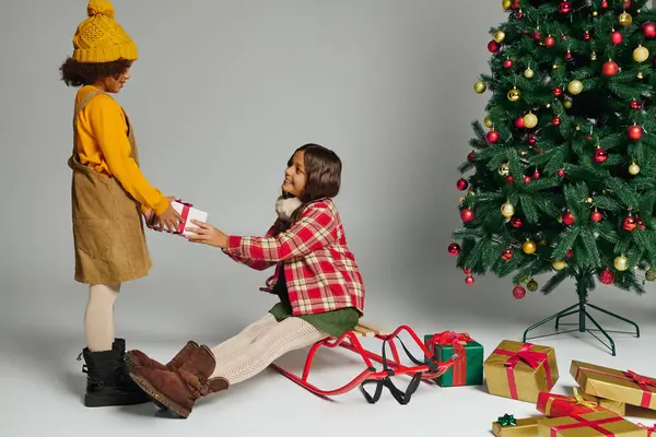 Two young girls share a delightful moment exchanging gifts by a beautifully decorated tree. — Stock Photo