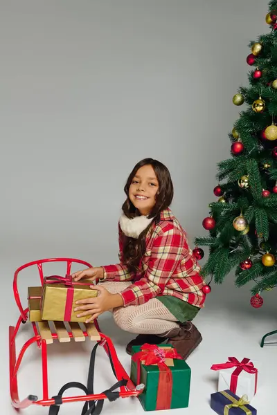 Una ragazza felice si siede con regali avvolti accanto a una classica slitta vicino a un albero di Natale decorato. — Foto stock