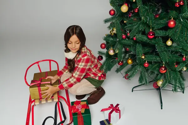 Una ragazza organizza felicemente regali su una slitta festiva accanto a un albero di Natale splendidamente decorato. — Foto stock