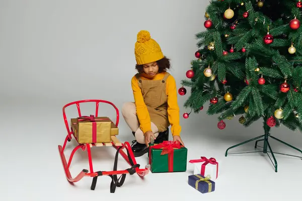Little girl in bright knit hat arranges colorful gifts near a decorated Christmas tree — Stock Photo