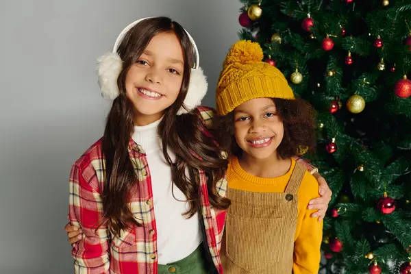 Two happy girls pose with bright smiles by a decorated Christmas tree, full of cheer. — Stock Photo