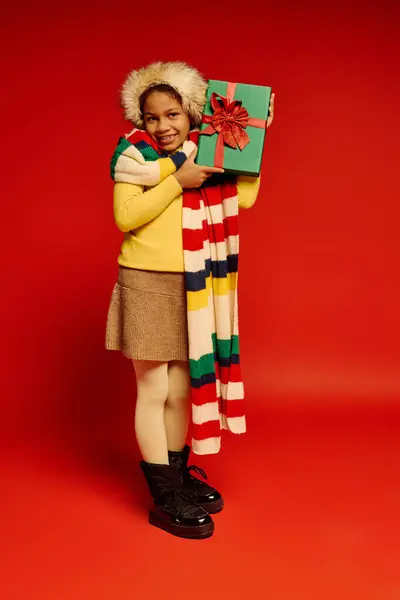Ragazza allegra vestita con abiti natalizi, con un regalo colorato e un sorriso luminoso. — Foto stock