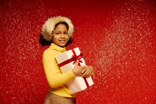 A smiling girl in a cozy sweater holds a gift, set against a bright red background. — Stock Photo