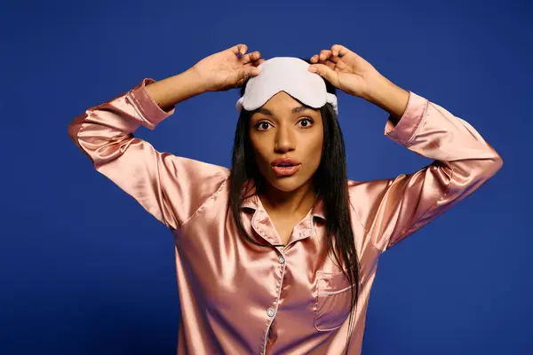 A stylish young African American woman wearing silk pajamas poses with a sleep mask in a studio. — Stock Photo