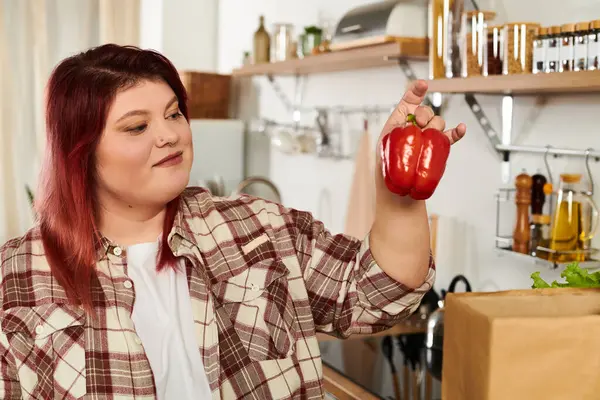 Giovane donna con più dimensioni figura sorride mentre tiene in mano un vivace peperone rosso nella sua cucina. — Foto stock