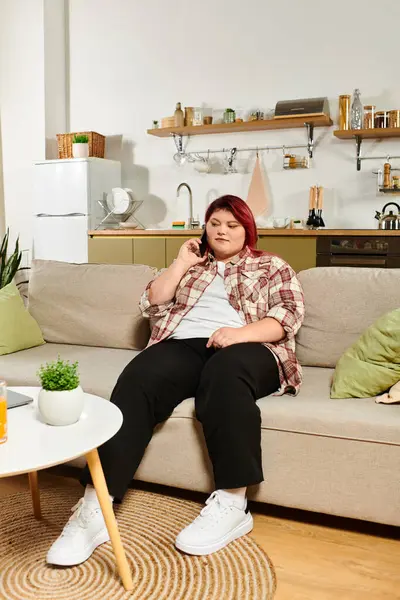 Joven mujer de talla grande disfruta de una llamada telefónica mientras descansa en un cómodo sofá. - foto de stock