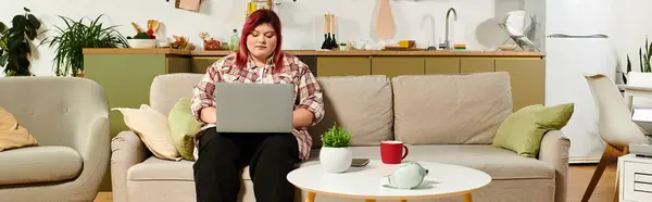 Jovem mulher plus size senta-se confortavelmente em um sofá digitando em seu laptop em uma cozinha moderna. — Fotografia de Stock