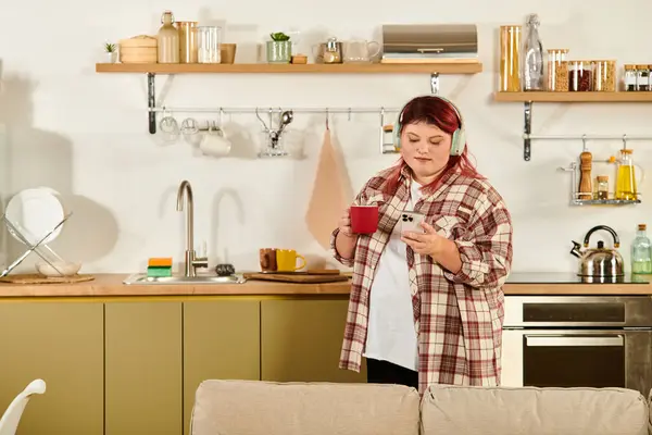 Una donna plus size gode il suo caffè mentre controlla il suo telefono in una cucina moderna. — Foto stock