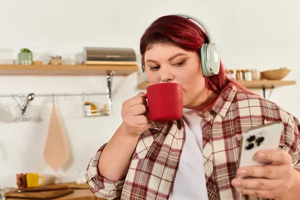 Plus size woman relishes a warm beverage while checking her phone in the kitchen. — Stock Photo