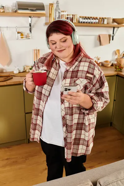 Una donna allegra plus size tiene una tazza mentre controlla il telefono in una cucina elegante. — Foto stock