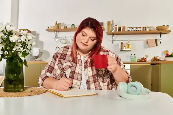 Женщина plus size расслабляется с теплым напитком, когда она творчески пишет в своем стильном кухонном пространстве. — стоковое фото