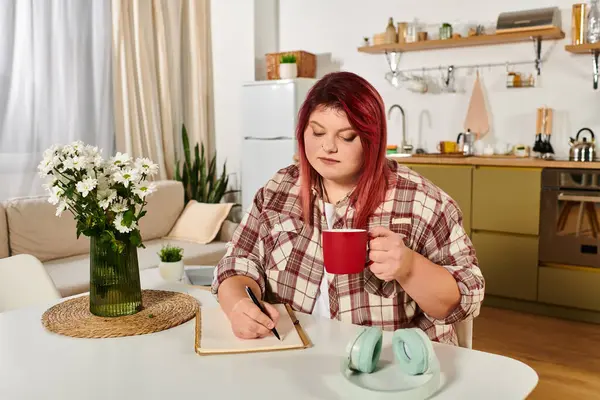 A beautiful young plus size woman writes thoughtfully while enjoying her coffee in a cozy kitchen. — Stock Photo