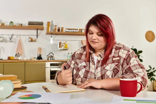 Beautiful plus size woman focuses on her notebook, creating ideas in a bright kitchen setting. — Stock Photo