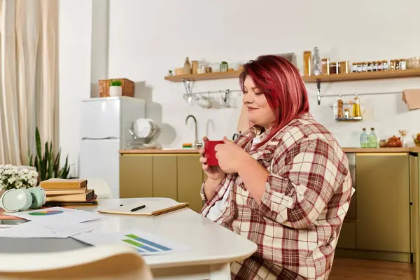 Una joven mujer de talla grande se sienta en una mesa disfrutando de una bebida caliente, rodeada de libros y notas. - foto de stock