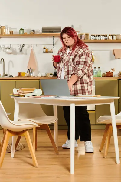 Una joven mujer de talla grande se para en una mesa, café en mano, enfocada en su trabajo. - foto de stock