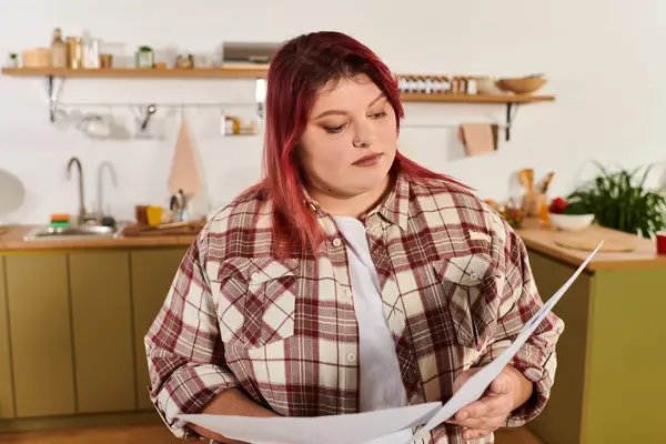 Brightly lit kitchen features a young plus size woman engaged in reviewing handwritten recipes. — Stock Photo