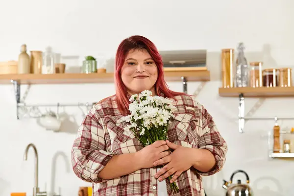 A plus size woman with bright hair holds a bouquet of flowers in a cozy kitchen atmosphere. — Stock Photo