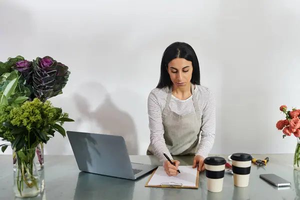 Un fleuriste concentré écrit des notes tout en étant entouré de fleurs fraîches et de tasses à café. — Photo de stock