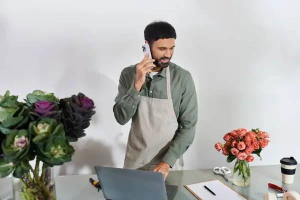 Un bell'uomo barbuto parla al telefono mentre organizza fiori freschi nel suo negozio. — Foto stock