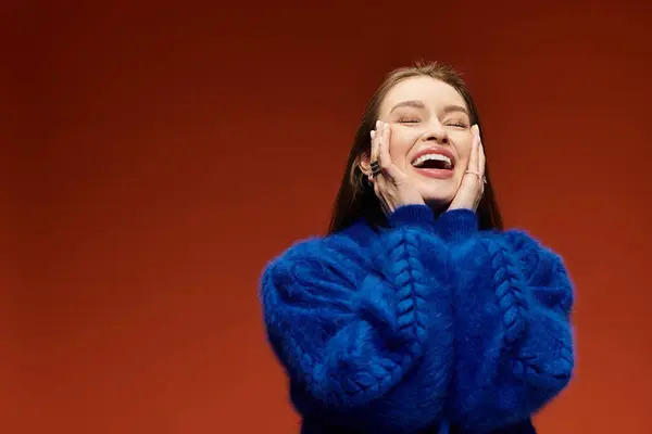 A joyful woman wearing a cozy sweater smiles widely while touching her face, exuding happiness. — Stock Photo