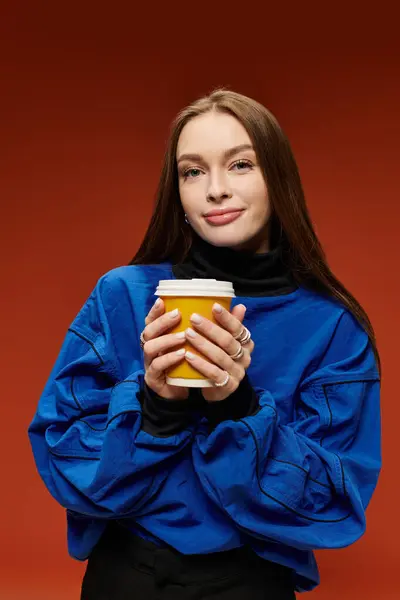 A vibrant woman in a blue sweater sips coffee against a warm orange backdrop. — Stock Photo
