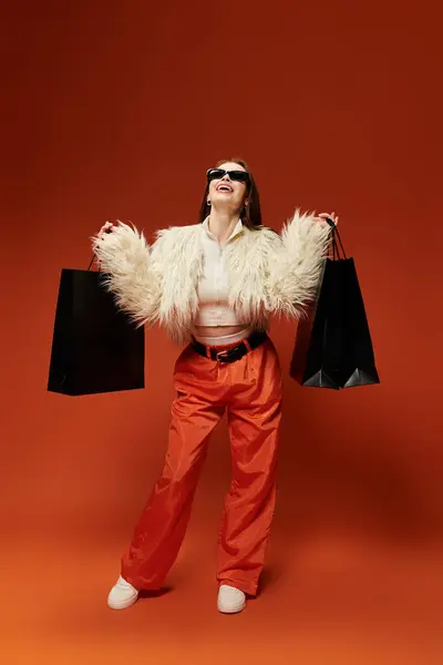 Woman joyfully holds shopping bags in a trendy outfit against a bright backdrop. — Stock Photo