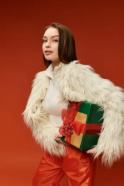 A beautiful woman in a furry coat presents a colorful gift box, embodying holiday cheer and style. — Stock Photo