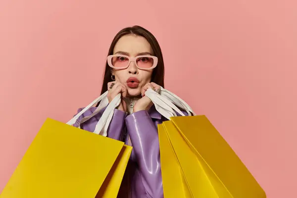 Una mujer elegante posa con coloridas bolsas de compras contra una pared rosa, haciendo pucheros juguetonamente. - foto de stock