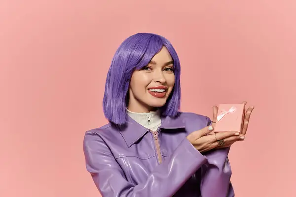 Una mujer elegante con el pelo morado sonríe alegremente, sosteniendo un producto contra un telón de fondo rosa. - foto de stock