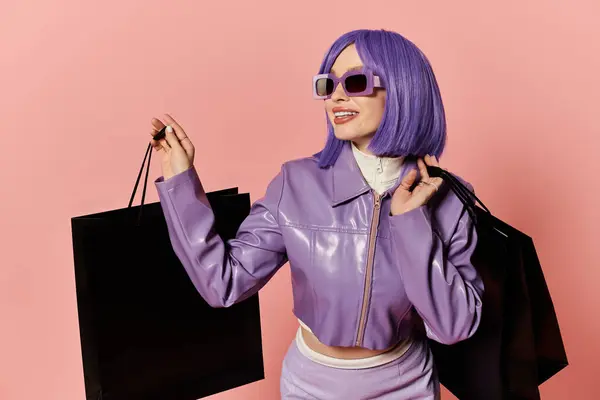 A stylish woman with purple hair smiles, holding black bags against a pink background. — Stock Photo