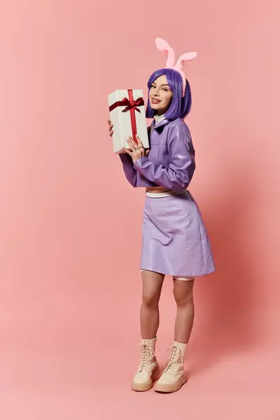 Fashionable woman stands in pink background, showcasing a gift box with playful bunny ears. — Stock Photo