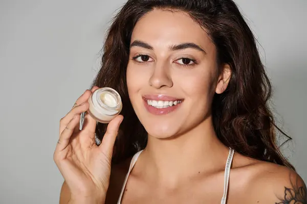 Beautiful young woman holds skincare cream, smiling confidently against a light backdrop. — Stock Photo
