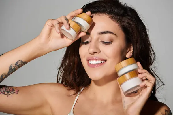 Beautiful young woman with a bright smile holds skincare jars against a neutral backdrop. — Stock Photo