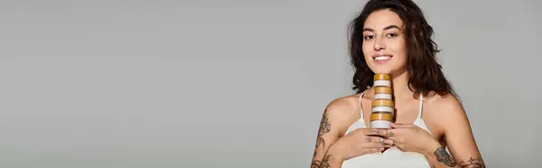 Giovane donna con tatuaggi sorride mentre tiene in mano vasi per la cura della pelle in un ambiente minimalista. — Foto stock