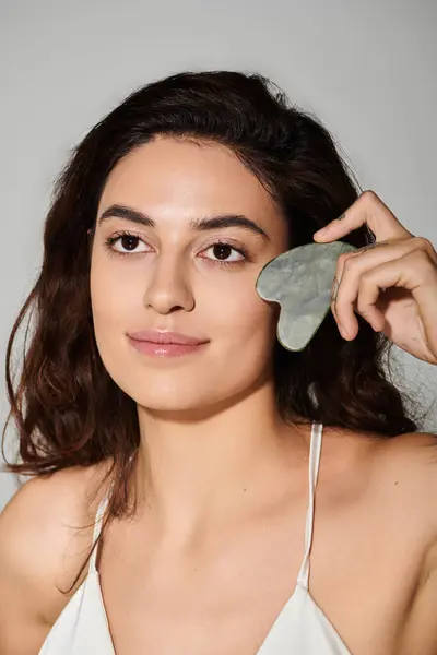 A beautiful young woman is holding a jade gua sha tool close to her face while relaxing indoors. — Stock Photo