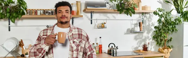 A young handsome man stands in a bright kitchen, holding a cup of coffee, surrounded by greenery. — Stock Photo