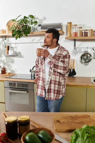 A young handsome man sips a warm drink in a stylish kitchen, surrounded by fresh produce. — Stock Photo