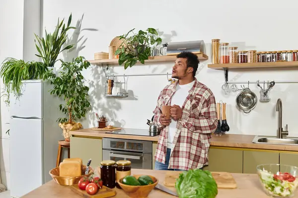 Man stands in a stylish kitchen, contemplating while surrounded by fresh vegetables and herbs. — Stock Photo