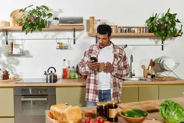 Handsome young man stands in a bright kitchen checking his phone during morning coffee. — Stock Photo
