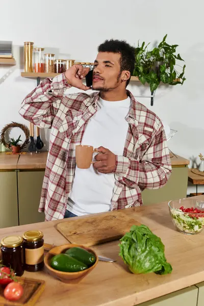 Young man enjoys a phone conversation in a well lit kitchen, holding a drink. — Stock Photo