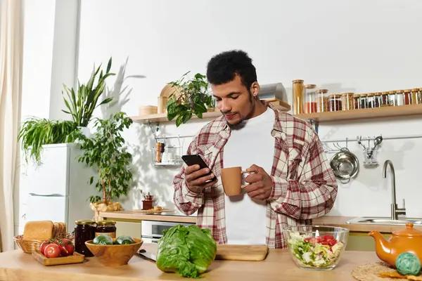 Schöner junger Mann steht in einer modernen Küche, nippt am Kaffee und surft auf seinem Smartphone. — Stockfoto