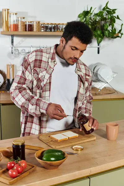 Handsome young man spreads jam on bread in a bright kitchen filled with ingredients. — Stock Photo